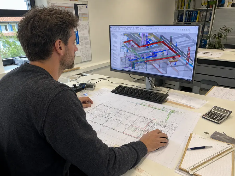 Fachplaner prüft am CAD-Arbeitsplatz das 3D-Modell der Gebäudetechnik neben dem gedruckten Grundrissplan
